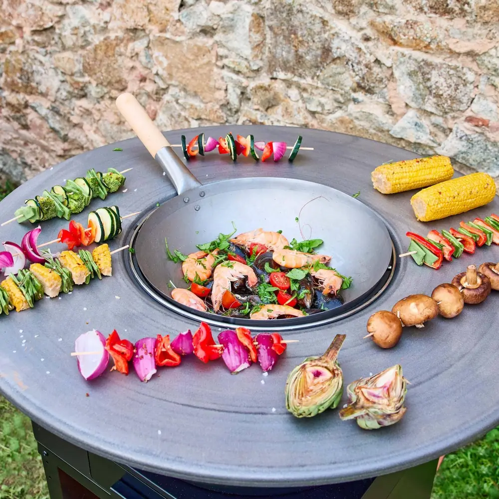 Brasero barbacoa Mombacho Ø 81,5 cm con plancha y mueble para leña: brochetas, mazorcas y setas en la plancha, con un salteado en el centro