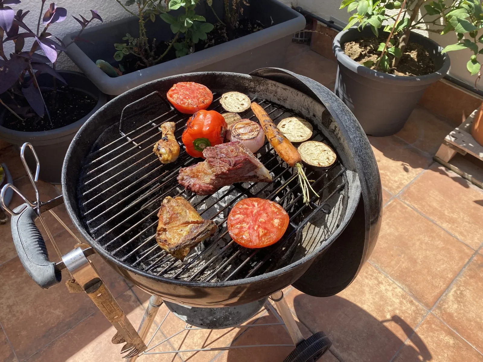 Barbacoa de carbón redonda con tapa en una terraza, con verduras y carne sobre la parrilla