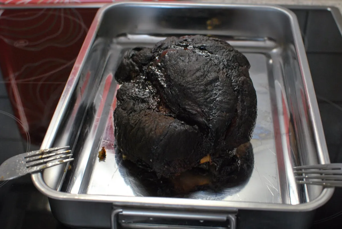 Paleta de cerdo ahumada con la corteza oscura en una bandeja de metal, lista para reposar antes de desmechar el pulled pork