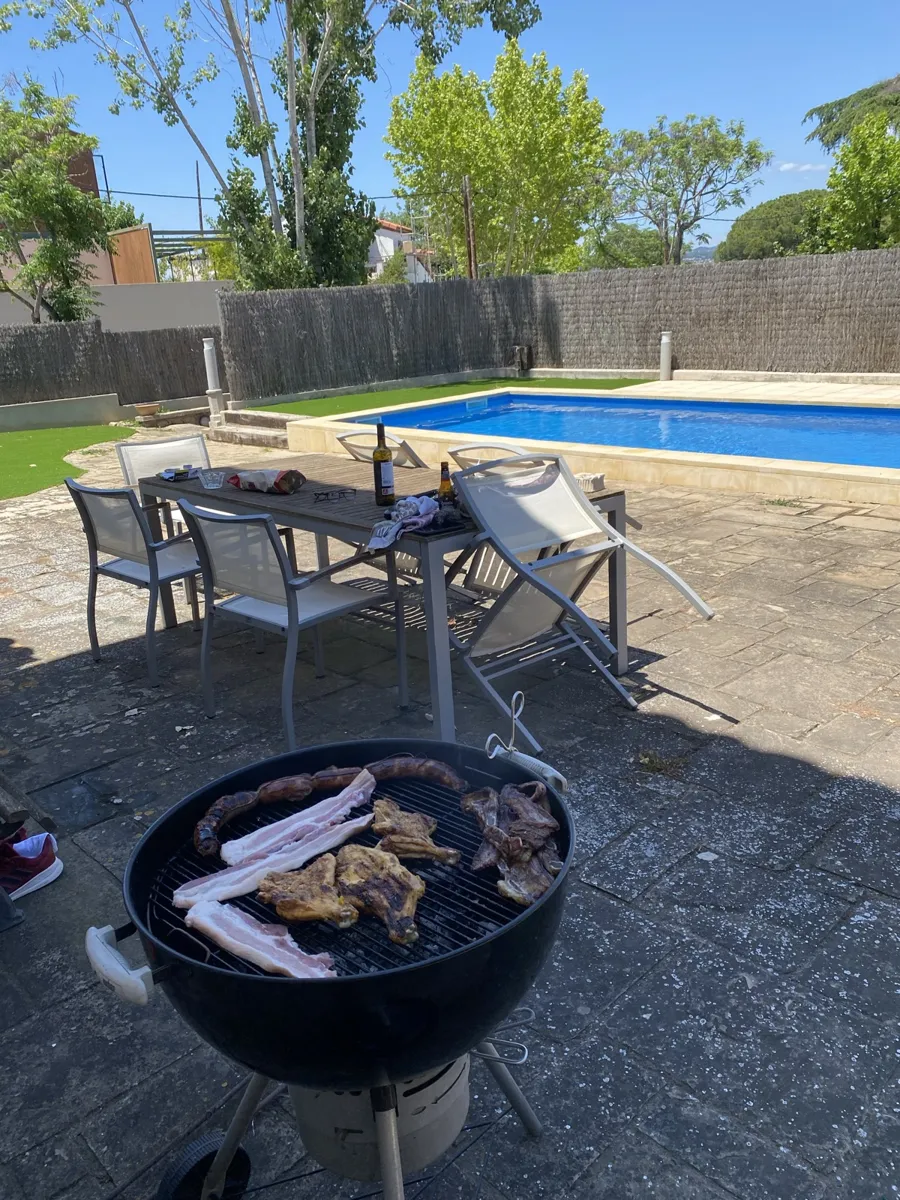 Barbacoa de carbón redonda con carne en la parrilla en un jardín con piscina y mesa de exterior